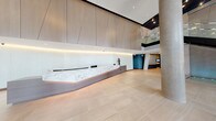 This expansive hotel lobby is designed with a refined contemporary aesthetic, combining high-end materials and architectural details to create a welcoming and impressive first impression. The space is anchored by a striking reception desk clad in veined white marble with warm under-counter lighting, set against a backdrop of floor-to-ceiling wood-paneled walls with subtle geometric detailing and recessed linear illumination.

The lobby's soaring double-height ceiling amplifies the sense of openness, while large-format neutral stone tiles extend across the floor, lending a clean and cohesive look throughout. Substantial cylindrical concrete columns punctuate the space, adding an industrial contrast to the otherwise polished finishes.

A grand staircase with a glass balustrade ascends along one side, accented by a dramatic water feature that catches light behind it. Pendant lighting with geometric sculptural elements hangs above a comfortable lounge area, furnished with contemporary sofas and chairs arranged around a low coffee table, ideal for guests to gather.

The entrance features floor-to-ceiling glazing and automatic sliding doors, flooding the lobby with natural light and providing open views of the surrounding landscape. A luggage cart near the entry reflects the space's full-service hospitality function.