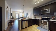 Located at 2421 Landmark Dr in Raleigh, NC, this single-floor residence offers a contemporary and well-designed living experience. The open-concept kitchen is a standout feature, boasting dark espresso cabinetry that extends to the ceiling, stainless steel appliances including a refrigerator, range, microwave, and dishwasher, and a granite-topped island with pendant lighting overhead. A textured tile backsplash and warm wood-tone flooring complete the polished aesthetic. The kitchen flows naturally into the living area, which features a ceiling-mounted track light fixture, large windows that invite natural light, and access to an outdoor area. The living room is generously proportioned, accommodating a large sectional sofa and a glass coffee table beneath a modern chandelier. The bedroom is a calm and comfortable retreat, featuring plush carpeting, a ceiling fan, and wide windows fitted with blinds and drapes that allow for adjustable natural light. The bathroom continues the dark cabinetry theme with a dual-sink vanity, granite countertops, wood-look flooring, and bold panel-style doors that add a refined, modern touch throughout. The overall design is cohesive, blending contemporary finishes with functional layouts.