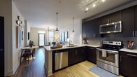 Located at 2421 Landmark Dr in Raleigh, NC, this single-floor residence offers a contemporary and well-designed living experience. The open-concept kitchen is a standout feature, boasting dark espresso cabinetry that extends to the ceiling, stainless steel appliances including a refrigerator, range, microwave, and dishwasher, and a granite-topped island with pendant lighting overhead. A textured tile backsplash and warm wood-tone flooring complete the polished aesthetic. The kitchen flows naturally into the living area, which features a ceiling-mounted track light fixture, large windows that invite natural light, and access to an outdoor area. The living room is generously proportioned, accommodating a large sectional sofa and a glass coffee table beneath a modern chandelier. The bedroom is a calm and comfortable retreat, featuring plush carpeting, a ceiling fan, and wide windows fitted with blinds and drapes that allow for adjustable natural light. The bathroom continues the dark cabinetry theme with a dual-sink vanity, granite countertops, wood-look flooring, and bold panel-style doors that add a refined, modern touch throughout. The overall design is cohesive, blending contemporary finishes with functional layouts.