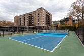 Located at 185 N West Temple in the heart of Salt Lake City, this outdoor amenity space offers a well-appointed recreational area designed for active use and social gathering. The centerpiece is a full-sized, dual-tone blue and green hard-surface court, lined and netted for both pickleball and tennis play. Tall sport lighting poles surround the court, ensuring usability during evening hours, while a chain-link and iron perimeter fence encloses the playing area for safety.

Adjacent to the court, a turf putting green provides a dedicated space for golf practice, complemented by nearby cornhole boards branded with the property's logo. A covered pergola seating area with outdoor lounge furniture offers a comfortable spot to relax between activities. Landscaping throughout the space features ornamental grasses, shrubs, and deciduous trees that provide seasonal color and a natural buffer from the surrounding environment.

The setting delivers striking panoramic views of the Salt Lake City skyline and the Wasatch mountain range in the distance, creating a dynamic urban backdrop. Covered parking canopies border a portion of the space, and the multi-story brick residential building rises prominently nearby. The overall design balances active recreation with comfortable outdoor living in a vibrant downtown setting.