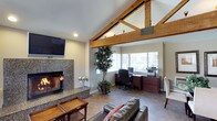 The clubhouse at 3634 College in Oceanside is a single-floor community space designed to offer both comfort and functionality. The interior is immediately distinguished by soaring vaulted ceilings adorned with prominent exposed wood beam trusses, lending the space a warm, lodge-inspired architectural character. Natural light flows in generously through large windows, brightening the open layout and providing views of the surrounding area. The main gathering area features a striking granite-surround fireplace, creating a cozy focal point ideal for relaxation and community gatherings. Leather seating is positioned to take full advantage of this inviting atmosphere. A wall-mounted flat-screen television adds a modern, entertainment-ready element to the space. A dedicated workstation area is thoughtfully integrated near the windows, complete with a desk, computer setup, and filing storage — offering residents a convenient space for productivity. Tile flooring runs throughout, providing durability and ease of maintenance. Decorative indoor plants add touches of greenery, softening the space and enhancing its overall ambiance. The combination of rustic architectural details, modern amenities, and natural light makes this clubhouse a versatile and welcoming community hub.