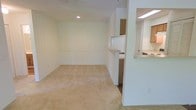 This single-floor space features an open-concept layout that seamlessly connects the living area to the kitchen. The living area is finished with light beige wall-to-wall carpeting, contributing to a warm and understated atmosphere. Neutral white walls run throughout, complemented by recessed lighting in the ceiling that provides focused illumination. A subtle chair rail detail lines the living area walls, adding a modest architectural accent.

The kitchen is positioned adjacent to the living space and is accessible via a pass-through counter, which also serves as a convenient bar-style surface. The kitchen features light wood cabinetry, a white refrigerator, and what appears to be a standard sink and countertop setup. An overhead fluorescent light fixture illuminates the kitchen workspace effectively. The pass-through opening enhances the sense of connectivity between the two areas, making the overall layout feel open and communicative despite its modest scale. The space presents a clean, move-in-ready aesthetic with a simple and practical design throughout.