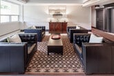 Located at 150 Allan St in Oakville, Ontario, this sophisticated lobby offers a refined and welcoming first impression. The space is designed with a classic aesthetic, combining clean architectural lines with timeless interior details. Crown moulding and a coffered ceiling add a sense of structure and elegance overhead, while large windows along one side flood the area with natural light and frame views of the surrounding greenery.

The central seating area features a set of tufted black leather chairs and sofas arranged around a dark rectangular coffee table, all anchored by a richly patterned area rug. A stately wooden credenza serves as a focal point along one wall, adorned with matching table lamps and a landscape artwork that enhances the refined atmosphere.

White tile flooring transitions seamlessly throughout the space, complemented by decorative area rugs near the entrance. A set of dark wood-panelled elevator doors adds a bold architectural contrast, while a well-lit corridor extends toward additional building amenities, including washroom facilities. The glassed entry vestibule provides a smooth transition from the outdoors, with a potted indoor tree near the windows adding a natural, organic touch to the polished interior.