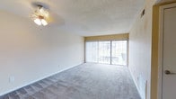 This single-floor space features an open and airy living area finished with wall-to-wall neutral carpeting that lends a clean, comfortable feel underfoot. The room is illuminated naturally by a large sliding glass door fitted with vertical blinds, which opens to an outdoor view and allows generous amounts of daylight to flow throughout the interior. A ceiling fan with an integrated light fixture is centrally mounted on the textured popcorn ceiling, providing both ambient lighting and air circulation. The walls are painted in a soft, warm neutral tone that complements the light carpeting and contributes to the room's bright, welcoming atmosphere. Clean white baseboards frame the perimeter of the space, adding a crisp, finished look. The layout is open and unobstructed, offering flexible possibilities for furniture arrangement. The sliding glass door provides a view of an exterior balcony railing and surrounding greenery, connecting the interior to the outdoors. Overall, the space presents a straightforward, functional design characterized by natural light, neutral finishes, and practical features suited for comfortable everyday living.