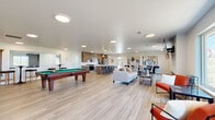 Located at 4451 Portrush Rd in Rapid City, South Dakota, this single-floor community space is thoughtfully designed to accommodate a variety of social and recreational activities. The open layout is unified by light wood-tone flooring that runs throughout, contributing to a clean, modern aesthetic complemented by white walls and flush-mount ceiling lighting.

The main gathering area features a comfortable lounge with a sectional sofa, accent chairs, a wall-mounted flat-screen television, and a sleek linear fireplace beneath it—creating a welcoming focal point. Large windows along the perimeter flood the space with natural light and provide views of the surrounding South Dakota landscape.

A dedicated game area includes a full-size pool table and a shuffleboard table, accompanied by bar-height seating and a pass-through window, making it well-suited for social events. Bold contemporary artwork adorns the walls throughout, adding visual character.

The fully equipped kitchen features warm wood cabinetry, stainless steel appliances including a French door refrigerator and range, a dishwasher, and a large island with marble-style countertops and bar seating illuminated by pendant lighting. A separate dining area with table seating completes the space, providing a functional area for group meals or gatherings.