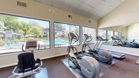 Located at 530 N Civic Dr in Walnut Creek, CA, this fitness center is a well-equipped exercise facility designed to support a complete workout routine. The single-room layout is thoughtfully organized, making efficient use of its space while maintaining an open, airy feel.

A defining architectural feature is the soaring vaulted ceiling on one end of the room, which lends a sense of openness and grandeur. Large, floor-to-ceiling windows line the main wall, flooding the interior with abundant natural light and offering unobstructed views of the outdoor pool area and surrounding landscaped grounds — creating an inviting and energizing atmosphere for workouts.

The cardio zone is well-stocked with Precor equipment, including elliptical trainers, treadmills, a recumbent bike, and a rowing machine, all positioned to take advantage of the pool views. The strength training area features a dumbbell rack with a flat bench, providing options for free weight exercises. Full-length mirrors on one wall enhance the sense of space and allow users to monitor their form.

The facility also includes a wall of smart Hub package lockers, adding a practical convenience feature for residents. The flooring throughout consists of durable carpet tiles, and the neutral, warm-toned walls contribute to the clean, functional aesthetic of the space. A glass entry door opens directly to the outdoor grounds, reinforcing the connection to the surrounding environment.