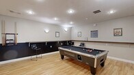Located at 517 Yosef Dr in Boone, NC, this dedicated game room is a well-appointed recreational space designed for social entertainment. The room features warm light hardwood flooring throughout, clean white walls accented by a dark wainscoting panel that runs along the lower portion of the walls, and a continuous wooden ledge that serves as a convenient shelf or resting surface.

The space is anchored by a full-size pool table with a dark felt surface and coin-operated design, positioned centrally to allow ample room for play. Adjacent to it, a regulation-size blue ping-pong table provides an additional competitive outlet. A wall-mounted flat-screen television is positioned on one wall, ideal for catching a game or streaming entertainment between matches.

Recessed ceiling lighting and wall-mounted sconces provide even, comfortable illumination throughout the room. Several bar-height chairs are distributed along the walls, offering casual seating for spectators. Decorative framed artwork and informational signage add personality to the space. An exit door connects directly to an adjacent fitness area, making this game room part of a broader amenity suite. The overall ambiance is clean, functional, and inviting — well-suited for community recreation.
