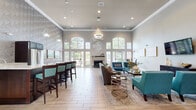 Located at 6690 Double Eagle Dr in Woodridge, Illinois, this single-floor community clubhouse is designed to impress with its open, airy layout and thoughtfully curated interiors. The main lounge area commands attention with soaring ceilings adorned with elegant crown molding and a combination of recessed lighting and statement pendant fixtures that cast a warm, inviting glow. A wall of arched windows and glass-paned French doors floods the space with natural light while offering views of the surrounding greenery. A stone-accented fireplace serves as a striking focal point, anchoring the seating area where plush sofas and vibrant teal accent chairs are arranged around gold-toned glass coffee tables. A bar-height counter with dark wood stools lines one side of the room, ideal for casual gatherings. Adjacent areas offer additional seating arrangements, including a wall-mounted TV lounge and high-top tables suited for work or conversation. A cozy nook near large windows provides an intimate two-seat table bathed in natural light, accented by curated artwork. Throughout, light wood-look tile flooring, geometric wallpaper accents, and a cohesive teal-and-neutral color palette create a polished, modern ambiance. Glass-paneled corridors connect to additional hallways, enhancing the sense of openness across this well-appointed community space.