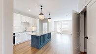 Located at 1140 Chambers Rd in Columbus, OH, this thoughtfully designed 2-bedroom, 2-bathroom residence offers a seamless blend of modern style and functional living across a single floor. The open-concept kitchen and living area serves as the heart of the home, showcasing crisp white cabinetry, a striking teal center island topped with granite countertops, a textured diamond-pattern tile backsplash, and a full suite of stainless steel appliances. Two globe pendant lights with matte black hardware add a sophisticated industrial touch overhead. Light wood-look flooring flows throughout the main living areas, enhancing the bright, airy atmosphere. A stylish barn door adds character to the hallway, which connects to both bedrooms. The primary bedroom features soft carpeting, dual windows for ample natural light, and an elegant wainscoting accent wall with gold-toned wall sconces. A spacious walk-in closet with multi-level shelving and hanging rods provides generous storage. The second bedroom is equally comfortable with plush carpet and natural light. The bathrooms are finished with granite-top white vanities, marble-look tile flooring, and a glass-enclosed walk-in shower with detailed tile surround and oil-rubbed bronze fixtures.