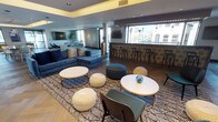 This contemporary amenity lounge is thoughtfully designed to offer a sophisticated and versatile gathering space across a single expansive floor. The interior is anchored by striking herringbone-patterned hardwood flooring that flows seamlessly throughout, complemented by tray ceilings with warm indirect lighting that add depth and elegance to the space.

The lounge is organized into several distinct zones. A prominent full-length bar with decorative tile cladding and a row of sleek black barstools serves as a central social hub, positioned in front of large windows that invite natural light and offer views of the surrounding exterior. Adjacent to the bar, a casual lounge area features plush blue sofas, round accent tables, woven pouf ottomans, and boldly patterned area rugs, creating an inviting atmosphere for relaxed conversation.

A separate seating nook is equipped with a large wall-mounted flat-screen television, a generous sectional sofa, and leather accent chairs arranged around a glass coffee table — ideal for media viewing or intimate gatherings. Toward the opposite end, an open dining area provides ample table and chair seating beneath recessed ceiling lights, with a console and display shelving along the walls.

Throughout the space, curated artwork, decorative accessories, and a cohesive navy, grey, and gold color palette lend a polished, upscale ambiance. Glass-paneled entry doors and multiple windows ensure the space remains bright and connected to the outside.