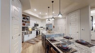 This single-floor residence offers a thoughtfully designed layout with high-quality finishes and a cohesive modern aesthetic. The kitchen is a standout feature, showcasing crisp white shaker cabinetry, open display shelving, a stainless steel farmhouse sink, stainless steel appliances, and a striking gray subway tile backsplash. A large granite-topped island with seating serves as the centerpiece, illuminated by a trio of industrial-style pendant lights and a tray ceiling overhead. Light wood-look flooring runs throughout, lending warmth and continuity to the space. The living area is bright and inviting, with large windows that allow natural light to fill the room, complemented by a statement pendant light fixture. A built-in desk nook with shelving provides a dedicated workspace tucked conveniently along a hallway. The bedrooms are comfortably sized, each with windows, soft neutral tones, and wood-look flooring. The primary bedroom features a warm accent wall and ample natural light. The bathroom is elegantly appointed with a granite vanity top, white shaker cabinetry, black hardware accents, a large framed mirror, wall sconces, and a walk-in glass-enclosed tile shower. Together, these spaces form a cohesive, well-finished residence suited for modern living.
