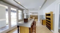 This single-floor community mail center is thoughtfully designed to blend functionality with a welcoming atmosphere. At the heart of the space is a long, gray-topped counter with elegant turned-leg detailing, lined with wooden bar stools that provide a comfortable area for sorting correspondence. Recessed lighting throughout the ceiling, complemented by modern pendant lamps, ensures the space is well-lit at all times. Large windows along one wall flood the interior with natural light, offering views of the surrounding landscaped exterior. An adjoining room, clearly marked "The Mail," houses a full wall of traditional brass individual mailboxes, serving as the operational hub of the center. To the side, warm wood cabinetry with a utility sink provides a practical work area, while additional shelving units offer organized storage. The light tile flooring and neutral gray-and-white color palette give the space a clean, polished look. Crown molding and tray ceiling details add an elevated architectural touch, lending the mail center a refined character that goes beyond purely utilitarian design.