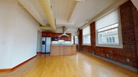 This single-floor space embodies classic urban loft design, seamlessly merging industrial elements with contemporary functionality. The open-concept living and kitchen area immediately captures attention with its striking exposed brick walls, large windows that invite abundant natural light and views of the surrounding cityscape, and prominent overhead ductwork that reinforces the space's industrial aesthetic. Light hardwood flooring runs throughout, enhancing the sense of openness and warmth.

The kitchen is well-appointed, featuring rich wood cabinetry, a central island with a butcher block countertop and built-in outlet, a stainless steel refrigerator, built-in microwave, dishwasher, and a statement overhead range hood. The layout positions the kitchen as an anchor of the open living area, ideal for both everyday use and entertaining.

Additional rooms, accessible from the main living space, include generously sized areas with continued hardwood flooring, exposed brick accent walls, built-in closets, and roller-shade windows. Two bathrooms feature tile flooring, wood vanities, and clean, functional layouts. The 3D floor plan reveals a well-organized, multi-room layout that balances open communal areas with more private spaces throughout.
