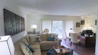 This single-floor space features an open-concept layout that seamlessly connects the living and dining areas, creating a cohesive and functional flow. The living area is anchored by a generously sized sectional sofa dressed in neutral tones and accented with teal and patterned throw pillows, complemented by a tufted ottoman that doubles as a coffee table. A large sliding glass door dominates the far wall, fitted with vertical blinds that diffuse natural light throughout the space while offering direct access to an outdoor deck or balcony. Views of surrounding trees are visible through the glass, lending a pleasant, nature-connected ambiance to the interior. The dining area sits adjacent to the living space, furnished with upholstered chairs around a dining table, maintaining the room's clean and coordinated aesthetic. Hardwood flooring runs through the dining area, adding warmth and contrast to the lighter tones of the living space. Wall-mounted artwork and tasteful décor accents contribute to the inviting atmosphere. The white walls and bright ceiling keep the interior feeling open and airy throughout.