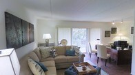 This single-floor space features an open-concept layout that seamlessly connects the living and dining areas, creating a cohesive and functional flow. The living area is anchored by a generously sized sectional sofa dressed in neutral tones and accented with teal and patterned throw pillows, complemented by a tufted ottoman that doubles as a coffee table. A large sliding glass door dominates the far wall, fitted with vertical blinds that diffuse natural light throughout the space while offering direct access to an outdoor deck or balcony. Views of surrounding trees are visible through the glass, lending a pleasant, nature-connected ambiance to the interior. The dining area sits adjacent to the living space, furnished with upholstered chairs around a dining table, maintaining the room's clean and coordinated aesthetic. Hardwood flooring runs through the dining area, adding warmth and contrast to the lighter tones of the living space. Wall-mounted artwork and tasteful décor accents contribute to the inviting atmosphere. The white walls and bright ceiling keep the interior feeling open and airy throughout.