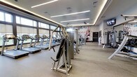 The fitness center at Station Village At Avenel is a thoughtfully designed, single-floor amenity space built to support a well-rounded workout experience. The room is generously proportioned, offering ample floor space for both cardio and strength training activities without feeling crowded.

A row of treadmills and cardio machines lines the windowed wall, bathing the workout area in natural light through large, floor-to-ceiling windows fitted with roller shades for adjustable brightness. Overhead, a tiered ceiling with recessed linear LED lighting lends a modern, polished aesthetic while ensuring the space is evenly and brightly illuminated throughout.

The strength training area features a selection of cable and weight machines arranged to allow easy movement between stations. Durable, rubber-backed flooring provides comfort and safety underfoot across the entire facility. Ceiling fans contribute to air circulation, keeping the environment comfortable during intense sessions.

A bold motivational mural anchors one wall, adding visual energy and reinforcing the active spirit of the space. The overall design balances functionality with a contemporary finish, creating a fitness environment that is both practical and visually appealing — a well-equipped amenity befitting the Station Village At Avenel community.