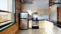 This single-floor space showcases an industrial loft aesthetic defined by a striking combination of exposed brick walls, heavy timber ceiling beams, and polished concrete floors throughout. The open layout is anchored by the main living area, where arched brick windows with black frames allow natural light to pour in, illuminating the raw architectural character of the space. A baseboard heater runs along the brick wall, and pendant lighting hangs from the timber-beamed ceiling, reinforcing the utilitarian design sensibility.

The kitchen is equipped with light wood cabinetry, dark countertops, stainless steel appliances including a refrigerator and electric range, and a central island with additional storage and sink. Exposed vertical pipes and overhead ductwork contribute to the industrial character of the space. The kitchen connects to the main living area via an open corridor framed by structural wooden columns.

A hallway off the main space leads to the bathroom, which features a pedestal sink, a mirrored medicine cabinet, and a full shower-tub combination. An entry door with an intercom system is positioned nearby, providing secure access. The overall ambiance is urban and raw, blending original building materials with functional modern amenities.