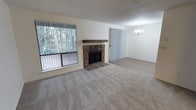 Located at 4950 Donovan Dr E in Olympia, Washington, this single-floor unit offers a comfortable and well-appointed layout designed for easy living. The open living and dining area serves as the heart of the home, anchored by a striking stone fireplace with a rustic wood mantel — a standout architectural feature that adds warmth and character to the space. Plush wall-to-wall carpeting runs throughout, contributing to a cozy and inviting atmosphere. Large windows and a sliding glass door flood the living area with natural light and frame peaceful views of the surrounding mature trees, creating a strong connection to the outdoors. Vertical blinds on both the windows and sliding door offer privacy and light control. The dining area is accentuated by an elegant chandelier, adding a touch of refinement to the space. A textured popcorn ceiling runs throughout the main living areas, a classic detail consistent with the building's era. The overall ambiance is calm and neutral, with a clean, move-in ready feel that provides a versatile canvas for personalization. This unit combines functional design with natural surroundings, making it a practical and pleasant place to call home in the heart of Olympia.