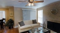 This single-floor space offers a welcoming and functional layout designed for comfortable everyday living. The living area serves as the heart of the home, finished in warm neutral tones that create a calm and inviting atmosphere. Crown molding adds a refined architectural detail along the ceiling line, while hardwood flooring grounds the space with a classic, durable finish. A ceiling fan with integrated lighting provides both air circulation and ambient illumination, contributing to the room's practical yet stylish character. Natural light filters in through multiple windows dressed with horizontal blinds and vertical panel blinds, offering flexible light control and a sense of openness. The living area provides ample space for seating and entertaining, with a layout that feels both organized and relaxed. The neutral wall tones pair seamlessly with the natural wood flooring, creating a cohesive palette that is easy to personalize. Overall, this space presents a clean, comfortable, and move-in ready environment suited for a variety of lifestyles, with thoughtful finishing touches that elevate the everyday living experience.
