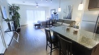This single-floor open-plan studio is thoughtfully designed to maximize functionality within a unified layout. Hardwood flooring extends throughout the entire space, lending warmth and visual continuity from the kitchen area through to the sleeping zone. A granite-topped breakfast bar anchors the kitchen side of the space, offering bar-height seating for two and serving as a natural divider between the kitchen and living areas. A pendant light hangs above the bar, providing focused task lighting that complements the overall illumination of the room.

The living area features a comfortable seating arrangement centered around an area rug, while a bookcase unit serves as a subtle visual separator between the lounge and sleeping zones without disrupting the open feel. The sleeping area is positioned toward the rear of the space near floor-to-ceiling vertical blinds that span the width of the wall, allowing generous natural light to fill the room. A ceiling fan is mounted overhead, contributing to both comfort and airflow. Track lighting along the ceiling provides additional, adjustable illumination. The overall ambiance is modern, clean, and efficiently organized.