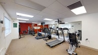 This fitness center is a dedicated workout facility designed to support a wide range of exercise routines. The space is outfitted with a selection of Vision Fitness cardio equipment, including treadmills, ellipticals, and a stationary bike, all arranged to allow comfortable movement throughout the room. Strength training machines are positioned along the far wall, offering users a variety of resistance and weight-based exercise options. A bold red accent wall adds a energizing visual contrast to the otherwise neutral color palette of white ceilings and light carpeted flooring. Full-length mirrors line one wall, enhancing the sense of space and allowing users to monitor their form during workouts. Overhead fluorescent lighting is integrated into a drop tile ceiling, providing bright, even illumination throughout the room. A wall-mounted television is positioned for visibility from the cardio equipment area, adding an entertainment option during workouts. A window on the far wall allows natural light to filter into the space, contributing to an open and airy atmosphere. The layout is thoughtfully organized to maximize usability while maintaining clear pathways between equipment stations.