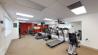 This fitness center is a dedicated workout facility designed to support a wide range of exercise routines. The space is outfitted with a selection of Vision Fitness cardio equipment, including treadmills, ellipticals, and a stationary bike, all arranged to allow comfortable movement throughout the room. Strength training machines are positioned along the far wall, offering users a variety of resistance and weight-based exercise options. A bold red accent wall adds a energizing visual contrast to the otherwise neutral color palette of white ceilings and light carpeted flooring. Full-length mirrors line one wall, enhancing the sense of space and allowing users to monitor their form during workouts. Overhead fluorescent lighting is integrated into a drop tile ceiling, providing bright, even illumination throughout the room. A wall-mounted television is positioned for visibility from the cardio equipment area, adding an entertainment option during workouts. A window on the far wall allows natural light to filter into the space, contributing to an open and airy atmosphere. The layout is thoughtfully organized to maximize usability while maintaining clear pathways between equipment stations.