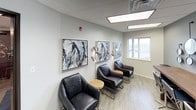 This well-appointed single-floor office space combines modern aesthetics with functional design, creating a polished and professional environment. The space is finished with light wood-look flooring throughout, drop tile ceilings with recessed panel lighting, and a neutral color palette of soft grays and warm whites that lend a calm, focused atmosphere.

A standout feature is the collaborative workspace anchored by a long, rustic solid-wood table paired with sleek navy blue bar-height chairs. The adjacent gray accent wall is adorned with an artful arrangement of varied circular mirrors, adding visual depth and a contemporary design element to the room.

The waiting or lounge area is thoughtfully curated with a set of modern leather armchairs arranged around small accent tables. Abstract artwork in muted tones lines the walls, while a floor lamp with adjustable spotlights provides ambient lighting. A window brings in natural light and offers a view of the surrounding exterior.

A dedicated utility nook houses office equipment such as a multifunction printer, supported by additional natural light from a window overlooking the outdoors. Interior glass-paneled doors with dark frames connect the various areas, maintaining an open, transparent feel while delineating functional zones throughout the space.