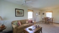 This single-story space features an open-concept layout connecting the living and dining areas. The living room is anchored by a plush, earth-toned sofa dressed with decorative pillows in warm red and striped patterns, complemented by a sturdy wooden coffee table at the center. A ceiling fan overhead provides both function and a finishing touch to the room. Natural light filters in through a window dressed with a patterned valance, brightening the soft, neutral-colored walls. Framed artwork adds visual interest, while a decorative branch arrangement in a floor vase contributes an organic, elegant accent. Neutral carpeting runs throughout, lending a warm and comfortable feel underfoot. The living area flows naturally into a dining space, which is furnished with bar-style seating — tall chairs positioned alongside a raised table — creating a casual yet defined eating area. Wall-mounted decorative accents, including a clock and additional framed pieces, add personality to the space. Overall, the interior presents a tidy, welcoming atmosphere with a cohesive color palette of warm neutrals and subtle earth tones throughout.