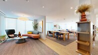 Located at 1001 N Coast Hwy in Oceanside, California, this thoughtfully designed single-floor space offers a generous and versatile layout spanning a comfortable footprint. The interior blends mid-century modern aesthetics with contemporary functionality, creating an inviting atmosphere suited for both socializing and focused work.

The living room serves as a welcoming lounge area, anchored by a sculptural curved sofa in warm coral and slate tones, complemented by bold patterned accent chairs, a round coffee table, and a large statement pendant light. Vibrant original artwork adorns the walls, and lush potted plants add a natural, refreshing element throughout. Floor-to-ceiling windows with vertical blinds flood the space with natural light and offer views of the surrounding coastal landscape, including glimpses of palm trees.

Two dining areas provide ample seating at long natural wood tables with upholstered chairs, ideal for group gatherings or collaborative work sessions. Built-in shelving units with curated décor and task lighting define additional lounge and work nooks throughout the space.

The space also includes dedicated conference rooms equipped with wall-mounted displays and seating for small groups, with one room offering direct views of the lush outdoor greenery. A well-appointed entry area with accent seating, a decorative mirror, and colorful artwork completes the welcoming atmosphere of this dynamic, amenity-rich space.