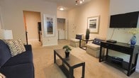 This single-floor residence offers an open and well-organized layout designed for comfortable everyday living. The living area is carpeted and generously proportioned, providing ample room for seating arrangements centered around a dark-finished coffee table. Neutral-toned walls and warm lighting from both a ceiling fixture and track lighting create a bright, inviting ambiance throughout.

A pair of upholstered armchairs with navy accent pillows anchor the seating area alongside sleek dark side tables, complemented by framed wall art that adds a polished touch. The high ceilings enhance the sense of openness and airiness within the space.

Visible through the living area, a dedicated laundry alcove houses an in-unit washer, offering added convenience. A white paneled door nearby leads to additional areas of the home. To the left, an open passageway connects to the kitchen, where black appliances and light cabinetry are visible, suggesting a functional and practical cooking space. The overall aesthetic is clean and neutral, making the space highly adaptable to a variety of furnishing styles.
