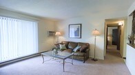This single-floor residence offers a comfortable and practical living environment with a clean, straightforward design. The living room serves as the heart of the space, furnished with a plush leather sofa accented by decorative throw pillows in muted earth tones. Soft wall-to-wall carpeting underfoot adds warmth and comfort to the room, while light-toned walls create a bright, airy atmosphere. A glass-top coffee table with an ornate metal base sits at the center of the seating area, providing both style and function. Two elegant floor lamps flank the room, casting warm ambient light throughout the space. A framed piece of abstract wall art adds a tasteful decorative touch above the sofa. Natural light filters in through a large window fitted with vertical blinds, enhancing the open feel of the room. A wall-mounted air conditioning unit ensures year-round climate comfort, while a baseboard heating unit along the wall provides an additional source of warmth. The textured ceiling adds a subtle architectural detail overhead. Overall, the space presents a tidy, move-in-ready interior with a neutral palette that lends itself well to a variety of personal styles and furnishing preferences.