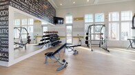 Located off I-75 in Sarasota, FL, this single-floor fitness center is designed to offer a bright, motivating environment for a full-spectrum workout experience. The space is characterized by soaring ceilings and an expansive wall of large, multi-pane windows that flood the interior with natural light and frame views of the surrounding palm-lined community.

The flooring throughout is light-toned hardwood, lending a clean and modern aesthetic. The walls feature a distinctive horizontal striped accent in gold and gray tones, complemented by an eye-catching typographic mural wall showcasing fitness-related words, adding an energetic visual element to the space.

The fitness center is comprehensively equipped across multiple zones. A dedicated strength training area houses a wide selection of selectorized cable machines and multi-functional resistance equipment. A free weights section provides a full dumbbell rack, adjustable benches, medicine balls, and stability balls, supported by wall-to-wall mirrors that maximize the sense of space. Mounted flat-screen televisions are positioned throughout for convenient viewing during workouts.

The cardio zone features treadmills with individual screens and a rowing machine, all positioned to take advantage of the sweeping outdoor views. Additional functional training accessories, including resistance bands and exercise mats, round out this thoroughly appointed fitness facility.