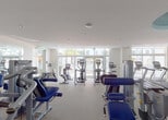 Located in Doral, Florida, this professional fitness center offers a well-appointed, single-floor layout designed to support a full range of workout routines. The space is generously sized, with high ceilings and large floor-to-ceiling windows that flood the interior with natural light and provide views of the outdoor pool area, creating an open and energizing atmosphere.

The cardio zone is equipped with multiple Matrix-brand treadmills, elliptical trainers, and stationary bikes, all featuring integrated digital displays. The strength training area is equally comprehensive, housing a multi-functional cable machine, a variety of selectorized resistance machines targeting major muscle groups, adjustable benches, and additional free-weight equipment. Full-length mirrors line the walls, enhancing the sense of space and allowing users to monitor their form throughout their workouts.

The interior design is clean and contemporary, with neutral tones accented by bold pops of yellow and a distinctive teal decorative column featuring colorful artwork, adding a vibrant visual focal point to the space. Durable, textured carpet flooring covers the workout areas, while the drop-tile ceiling incorporates recessed lighting to ensure consistent illumination throughout. The facility presents a polished, well-maintained environment suitable for a wide range of fitness needs.