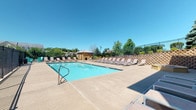 Located at 3101 Old 63 S in Columbia, Missouri, this outdoor pool facility serves as a well-appointed community amenity designed for relaxation and recreation. The centerpiece is a large, rectangular swimming pool filled with clear blue water, surrounded by an expansive concrete sun deck that provides ample room for lounging and movement. Dozens of slatted chaise lounge chairs are arranged along multiple sides of the pool, offering plentiful seating for residents to enjoy the open sky and sunshine. A hot tub is situated adjacent to the main pool, providing an additional relaxation option. The facility is enclosed by a sturdy black iron fence, with a controlled-access gate ensuring security and privacy for authorized users. A small pool house structure with light-colored siding is positioned within the enclosure, offering a convenient functional support area. Tiered retaining walls with decorative block work line one side of the deck, adding structured landscaping depth to the setting. Mature trees and shrubs are visible beyond the fence line, contributing a natural backdrop. Posted pool rules and safety signage are displayed prominently on the building exterior. The entire space benefits from abundant natural light and open views of the surrounding residential neighborhood under wide Missouri skies.