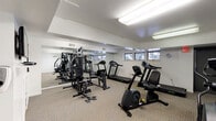 This fitness room offers a comprehensive workout environment designed to accommodate a variety of exercise routines. The space is laid out on a single level and finished with neutral carpet flooring throughout, creating a practical and comfortable surface underfoot. Bright fluorescent overhead lighting illuminates the entire room evenly, ensuring excellent visibility during workouts.

A full wall of mirrors lines one side of the room, visually expanding the space and allowing users to monitor their form during exercise. A row of horizontal windows along the upper portion of the walls allows natural light to filter in, offering views of surrounding greenery and contributing to a pleasant atmosphere.

The room is equipped with a multi-station cable strength trainer, multiple treadmills, an elliptical cross-trainer, a recumbent exercise bike, and a rowing-style machine, providing options for both cardiovascular and strength training. A wall-mounted flat-screen television adds an entertainment element for users during workouts. An exercise reference chart is posted on the wall near the strength equipment for guidance.

A glass-paneled door provides access to an adjacent area, and the clean white walls and organized layout give the space a tidy, functional character throughout.