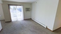 This single-floor space presents a straightforward and functional layout centered around a carpeted main living area. The room features neutral-toned walls and beige carpet that contribute to a clean, understated aesthetic throughout. A sliding glass door fitted with vertical blinds serves as a prominent architectural element along one wall, allowing natural light to filter into the interior while providing access to an outdoor area. Views through the glass door reveal an exterior railing and greenery beyond, adding a sense of openness to the space. Climate control is well-addressed, with a wall-mounted air conditioning unit installed above the sliding door and a baseboard heater running along the adjacent wall, ensuring year-round comfort. The room's layout is open and unobstructed, offering flexibility for various furniture arrangements. Electrical outlets are positioned conveniently along the baseboard, supporting practical use of the space. The overall ambiance is simple and move-in ready, with a functional design that prioritizes comfort and usability over decorative complexity.
