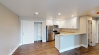 This single-floor, 2-bedroom, 2-bathroom residence offers a clean, modern aesthetic throughout. The open-concept kitchen serves as a central feature, showcasing crisp white shaker-style cabinetry that extends to the upper walls, providing generous storage space. Light-toned countertops complement the cabinetry, while a peninsula offers additional prep space and casual seating potential. Stainless steel appliances — including a refrigerator, microwave, and range — lend a contemporary, polished look to the kitchen area. Recessed lighting is installed throughout, ensuring consistent, well-distributed illumination across the space. Dark-toned hardwood-style flooring runs throughout, creating a striking contrast against the white cabinetry and light walls. A built-in closet with open shelving is conveniently positioned adjacent to the kitchen, offering versatile storage options. The neutral color palette of white and warm gray tones contributes to a bright, airy atmosphere throughout the unit. Overall, the space balances functional design with modern finishes, making it a practical and visually appealing single-level home.