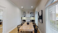 Located at 5750 Curtis Clark Dr in Corpus Christi, Texas, this single-floor space presents a well-designed, multifunctional room that seamlessly blends a dining area with a dedicated workspace. The room is finished with warm light hardwood flooring throughout, complemented by crisp white walls and crown molding that lend a clean, polished aesthetic.

A centerpiece of the space is the built-in workstation along one wall, featuring an extended white desk surface with under-cabinet lighting, dual seating positions, and generous overhead shelving with integrated cabinetry. This built-in unit provides ample storage and a functional work environment. Adjacent to the dining table, a rustic wood table surrounded by black Windsor-style chairs serves as an inviting gathering area.

Natural light is a defining feature, with prominent arched windows fitted with white horizontal blinds offering views of the surrounding greenery. Recessed ceiling lighting ensures the space remains bright and well-lit at all times. A glass-paned French door connects the room to an adjacent area, enhancing the sense of openness.

The space is accented with bold black-and-white abstract artwork, a neutral accent chair tucked into the corner, and thoughtfully placed décor on the shelving, creating an ambiance that is both contemporary and functional.