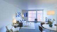 This single-floor space offers a thoughtfully designed open-plan layout that maximizes both comfort and functionality. The living area is the centerpiece of the unit, bathed in natural light streaming through expansive floor-to-ceiling windows that frame views of the surrounding urban streetscape. The clean, smooth white ceilings enhance the sense of openness and airiness throughout. The living space comfortably accommodates a contemporary seating arrangement, complete with a sofa, accent chairs, and a coffee table set atop a striped area rug that adds a subtle pop of color to the neutral palette. Soft, ambient lighting from floor lamps complements the natural light, creating a warm and inviting atmosphere. Colorful wall art provides visual interest against the soft gray tones of the walls. Adjacent to the living area, a compact dining nook features a round white table with modern curved chairs, offering a defined yet seamlessly integrated space for meals. The overall aesthetic is modern and minimalist, with a cool, neutral color scheme of grays and whites accented by touches of blue and green. The large windows remain a standout architectural feature, connecting the interior to the vibrant cityscape beyond.