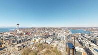 This rooftop terrace occupies the uppermost level of a prominent high-rise tower in Seattle, delivering unobstructed 360-degree views encompassing the city skyline, Puget Sound, surrounding neighborhoods, and the distant Olympic Mountains. The curved architectural form of the terrace is a defining feature, with its sweeping arc-shaped layout following the contours of the building's exterior.

The outdoor space is finished with large-format porcelain or concrete pavers underfoot, providing a clean, contemporary surface throughout. Glass panel railings, supported by sleek metal posts, line the perimeter, ensuring uninterrupted sightlines while maintaining safety at this significant elevation. The transparent barriers allow panoramic views from virtually every vantage point on the terrace.

Thoughtfully arranged seating areas are distributed across the terrace, featuring modular sectional sofas in neutral tones, complemented by planter boxes filled with ornamental grasses that add greenery to the urban setting. A prominent circular concrete fire pit serves as a central gathering focal point in one seating zone, enhancing the space's functionality across seasons.

Floor-to-ceiling glass curtain walls connect the indoor and outdoor areas, flooding any adjacent interior with natural light and maintaining visual continuity with the dramatic cityscape beyond. The overall ambiance is sleek, contemporary, and distinctly urban.