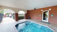 Located in Canton, Mississippi, this single-level covered pool facility offers a well-appointed aquatic retreat designed for comfort and functionality. The pool area is framed by classic red brick walls that lend a warm, durable character to the space, complemented by a decorative arched entryway that adds an elegant architectural touch. A ceiling fan is mounted overhead to promote airflow and keep the covered area comfortable during warmer months. The pool itself features clean, blue water and is bordered by tiled coping that ties seamlessly into the surrounding hardscape. Wrought iron fencing encloses the perimeter, providing security while maintaining an open, airy feel with views of the surrounding greenery and landscaping beyond. A gate within the fencing offers convenient access, and signage is posted for pool rules and safety information. A door on the adjacent brick wall connects the pool area to an interior space, adding ease of access for guests or residents. The covered ceiling overhead, finished in white, keeps the area shaded and protected from the elements, making this an inviting all-weather outdoor amenity.