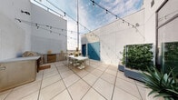 Located at 3921 Wilshire Boulevard in the vibrant Koreatown neighborhood of Los Angeles, this rooftop terrace offers a well-appointed open-air amenity space with striking views of the surrounding cityscape. The terrace is finished with large-format, light-toned porcelain tiles that lend a clean, modern aesthetic to the expansive floor area.

A dedicated outdoor dining zone anchors the space, featuring a white slatted dining table surrounded by coordinating metal chairs — ideal for gatherings and al fresco meals. Overhead, a canopy of bistro string lights crisscrosses the terrace, creating an inviting ambiance during evening hours. An outdoor kitchen station with stainless steel cabinetry and utility connections is positioned along one wall, providing functional support for entertaining.

Architectural interest is added by crisp white perimeter walls and a pair of bold teal double doors, which serve as a distinctive visual focal point. Planters with lush greenery, including a vertical garden element, introduce natural texture and life to the hardscape setting. Access to the terrace is provided through a floor-to-ceiling glass entry with ADA-compliant automatic door activation. The open sky above allows for abundant natural light throughout the day, making this rooftop terrace a versatile and stylish outdoor destination.