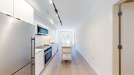 This single-floor unit showcases a clean, contemporary design throughout. The open-concept kitchen runs along one wall and is outfitted with flat-front white cabinetry, dark quartz countertops, a white tile backsplash, and a full suite of stainless steel appliances including a built-in oven, microwave, dishwasher, and refrigerator. Black track lighting overhead provides focused task illumination, while a movable kitchen island on casters adds flexible prep and storage space. Light-toned hardwood flooring extends throughout the main living areas, lending warmth to the otherwise bright, white interior.

At the far end of the unit, floor-to-ceiling windows and a glass door draw in generous natural light and provide views of the surrounding urban streetscape. The bedroom is a well-proportioned, quiet space with large windows overlooking neighboring architecture and ample room for furnishings.

The bathroom is thoughtfully designed with a long wood-toned vanity, white quartz countertop, a wide mirror with wall-mounted lighting, large-format tile flooring, and a glass-enclosed shower. A stacked washer and dryer unit is conveniently integrated into the bathroom space. The overall ambiance is modern, minimal, and move-in ready.