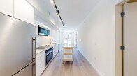This single-floor unit showcases a clean, contemporary design throughout. The open-concept kitchen runs along one wall and is outfitted with flat-front white cabinetry, dark quartz countertops, a white tile backsplash, and a full suite of stainless steel appliances including a built-in oven, microwave, dishwasher, and refrigerator. Black track lighting overhead provides focused task illumination, while a movable kitchen island on casters adds flexible prep and storage space. Light-toned hardwood flooring extends throughout the main living areas, lending warmth to the otherwise bright, white interior.

At the far end of the unit, floor-to-ceiling windows and a glass door draw in generous natural light and provide views of the surrounding urban streetscape. The bedroom is a well-proportioned, quiet space with large windows overlooking neighboring architecture and ample room for furnishings.

The bathroom is thoughtfully designed with a long wood-toned vanity, white quartz countertop, a wide mirror with wall-mounted lighting, large-format tile flooring, and a glass-enclosed shower. A stacked washer and dryer unit is conveniently integrated into the bathroom space. The overall ambiance is modern, minimal, and move-in ready.