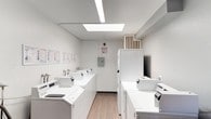 This shared laundry facility is a single-floor space designed with functionality and convenience in mind. The main laundry area is bright and clean, illuminated by recessed fluorescent lighting and a skylight that enhances the open feel of the room. A row of top-load washing machines lines one wall, accompanied by a set of stackable dryer units, providing ample capacity for multiple users. Light wood-style flooring runs throughout this section, contributing to the room's clean, well-maintained appearance. Instructional posted signage and washing guidelines are displayed along the walls for user reference, and a wall-mounted electrical panel ensures organized utility management. An HVAC unit is positioned overhead for climate control. The adjacent area features dark wood-style flooring and serves as a transitional and folding zone, complete with a laminate-topped counter for sorting and folding laundry. A Coca-Cola vending machine offers refreshments, making extended laundry visits more comfortable. A metal waste bin and a wall-mounted control panel are also present. The space is accessed through a vented utility door equipped with a fire extinguisher nearby, underscoring the facility's attention to safety. Overall, this laundry facility is a practical, thoughtfully arranged communal space built to serve a shared residential or hospitality setting efficiently.