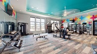 Located in Colorado Springs, CO, this fitness center offers a comprehensive workout facility designed with both functionality and aesthetics in mind. The space is defined by its eye-catching tray ceiling painted in a bold teal, accented by recessed lighting and a ceiling fan, creating an energizing atmosphere throughout. A vibrant wall mural featuring multicolored geometric cross shapes adds a lively, modern character to the interior, complemented by full-length mirrors that enhance the sense of openness.

The facility is comprehensively equipped to support a wide range of fitness routines. The strength training area includes a cable crossover machine, adjustable benches, a dumbbell rack, and various selectorized weight machines. The cardio section features StairMaster climbers, stationary bikes with digital displays, and elliptical trainers, all positioned along large windows that invite generous natural light and offer views of the surrounding Colorado Springs neighborhood.

The flooring consists of patterned modular carpet tiles in neutral tones with teal accents, tying together the facility's color palette. The tall windows along multiple walls ensure the space remains bright and connected to the outdoors. The overall layout is open and well-organized, providing ample room to move freely between equipment zones while maintaining a motivating and professional fitness environment.
