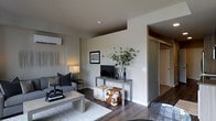 This single-floor residence showcases a thoughtfully designed interior with a clean, contemporary aesthetic. The living area serves as the heart of the space, featuring rich, dark hardwood flooring that adds warmth and contrast to the neutral, light-toned walls. A sleek wall-mounted air conditioning unit ensures year-round comfort, while the open layout creates a sense of spaciousness and ease of movement.

The living room is styled with modern furnishings, including a low-profile sofa with dark accent pillows and a minimalist coffee table atop a patterned area rug. A slender metal media console holds a flat-screen television, accompanied by a cowhide ottoman that introduces a touch of organic texture to the refined interior. A tall, leafy plant in a glass vase adds a fresh, natural element to the room.

A glass sliding door is visible in the background, leading to what appears to be a closet or storage area, reflecting a practical approach to space utilization. The overall ambiance is calm and modern, with muted tones, clean lines, and carefully chosen furnishings that work together to create a cohesive and inviting living environment.