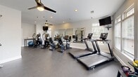 Located in Greer, South Carolina, this fitness center is a thoughtfully designed workout facility that offers a comprehensive range of exercise options within a clean, open layout. The space features durable rubber flooring throughout, providing a safe and functional surface suited for high-traffic athletic use. Large windows along one wall flood the room with abundant natural light, creating an energizing atmosphere, while a glass-paned entry door adds additional brightness and a welcoming feel. The cardio area is well-appointed with multiple treadmills, elliptical trainers, stair climbers, and a recumbent bike from the TRUE fitness brand, all arranged to maximize space and usability. Wall-mounted televisions and ceiling fans with lights enhance comfort during workouts. A large wall mirror spans one section of the room, allowing users to monitor their form while exercising. The free weight area features a full rack of dumbbells and a selection of kettlebells in varying sizes, complemented by a dedicated bench for strength training. A professional-grade cable machine rounds out the strength equipment offerings. Recessed lighting and ventilation fixtures are integrated into the ceiling, maintaining a comfortable and well-lit environment throughout the facility.