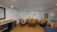 This single-floor space serves as a stylish and comfortable lounge area, designed with a clean, modern aesthetic. The room is anchored by a coordinated wicker furniture suite, including a sofa, an armchair, and a chair-and-ottoman pairing, all upholstered in striking orange cushions with gold accent pillows that add warmth and depth to the space.

The flooring throughout is a warm hardwood-style surface that complements the furniture tones and contributes to the inviting atmosphere. Recessed lighting in the white ceiling provides even, practical illumination across the room, while a central HVAC vent ensures year-round comfort.

A decorative wall panel, featuring circular botanical-inspired artwork in neutral tones, serves as a focal point behind the sofa, adding a subtle artistic element to the otherwise minimalist walls. A low, dark coffee table sits at the center of the seating arrangement, completing the conversation area.

Along one wall, a compact countertop with an under-counter refrigerator suggests a convenient refreshment station, making the space well-suited for social gatherings or communal use. Overall, the room balances functionality with contemporary design.