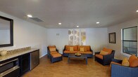 This single-floor space serves as a stylish and comfortable lounge area, designed with a clean, modern aesthetic. The room is anchored by a coordinated wicker furniture suite, including a sofa, an armchair, and a chair-and-ottoman pairing, all upholstered in striking orange cushions with gold accent pillows that add warmth and depth to the space.

The flooring throughout is a warm hardwood-style surface that complements the furniture tones and contributes to the inviting atmosphere. Recessed lighting in the white ceiling provides even, practical illumination across the room, while a central HVAC vent ensures year-round comfort.

A decorative wall panel, featuring circular botanical-inspired artwork in neutral tones, serves as a focal point behind the sofa, adding a subtle artistic element to the otherwise minimalist walls. A low, dark coffee table sits at the center of the seating arrangement, completing the conversation area.

Along one wall, a compact countertop with an under-counter refrigerator suggests a convenient refreshment station, making the space well-suited for social gatherings or communal use. Overall, the room balances functionality with contemporary design.